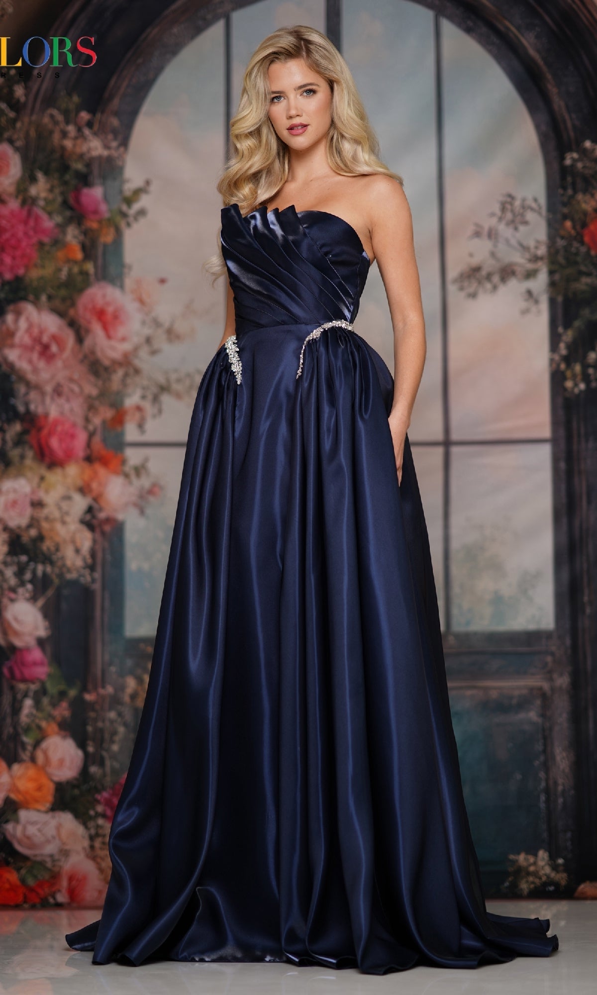 A woman wears the Strapless A-Line Prom Ball Gown: Colors Dress 3845 in dark blue, standing before a floral and arched window backdrop.